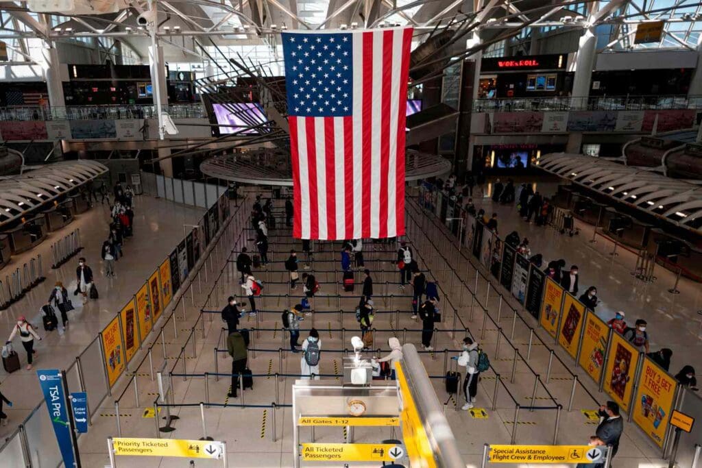 Large American flag hangs from the ceiling of a busy airport terminal with travelers lined up at security checkpoints below and bright yellow signage around the gates.